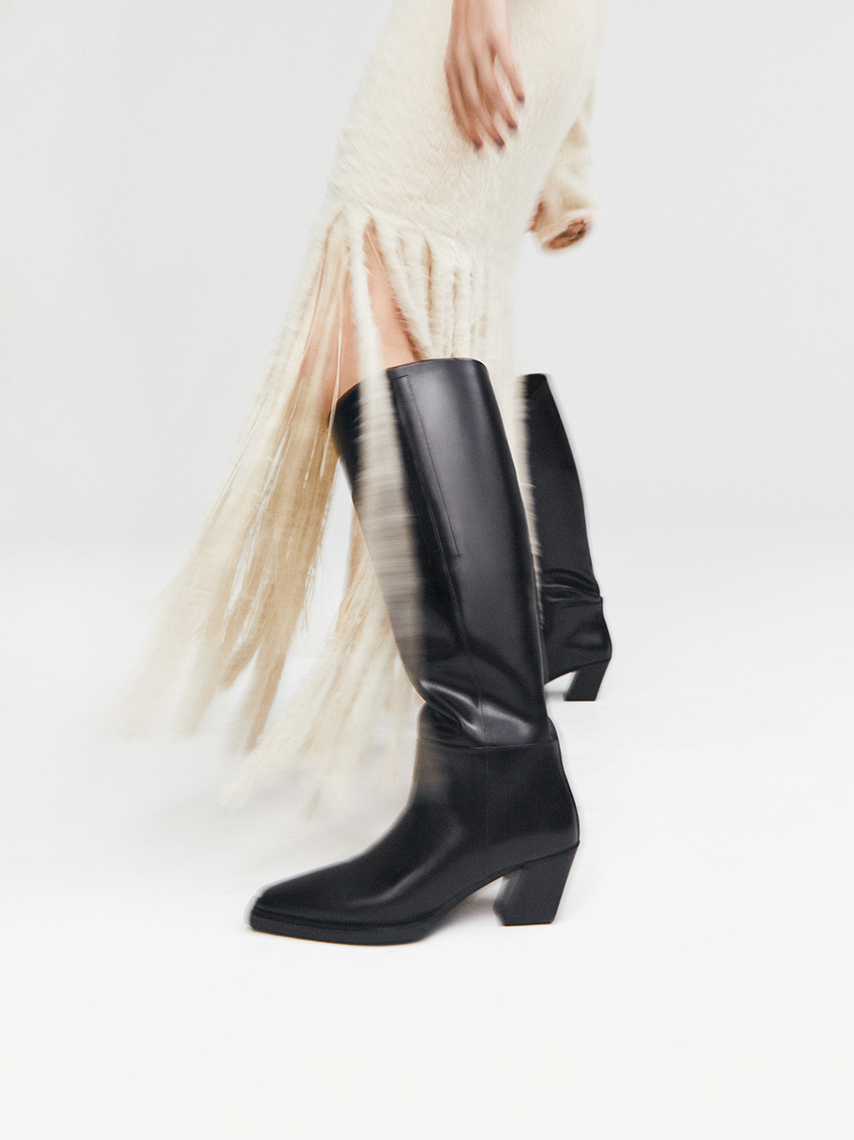 Knee-high western boots Alina in all-black, styled with a white fringe midi-dress.