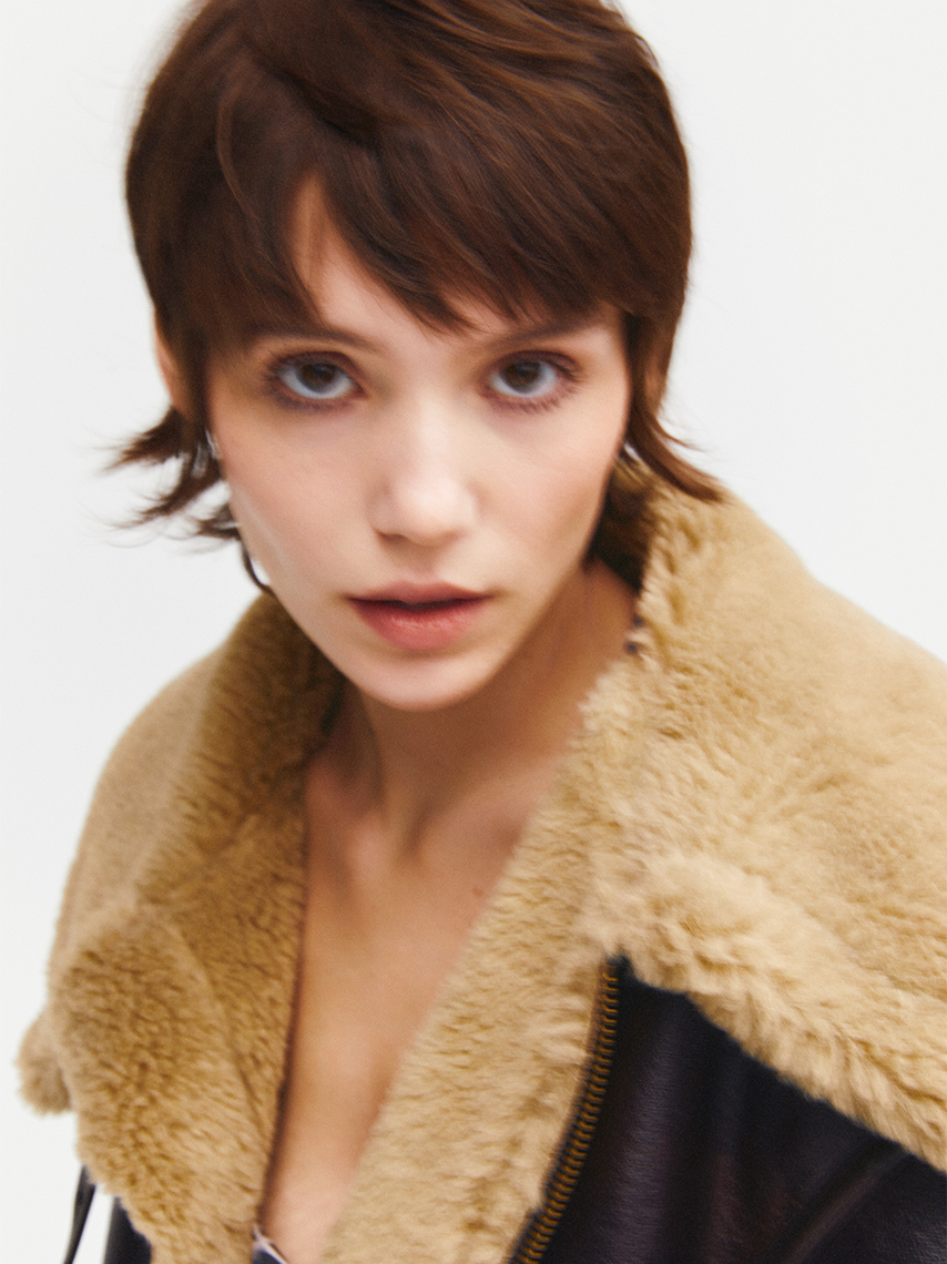 A portrait of a model with short, brown hair, wearing a black jacket with a beige collar.