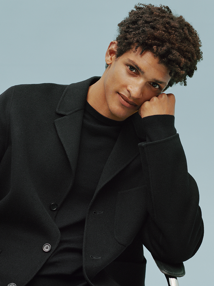 A portrait of a smiling model with brown, curly hair, wearing all-black clothing.