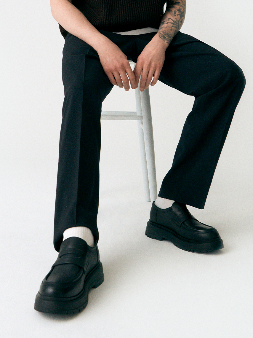 Den taturerade modellen sitter på en pall med svarta kostymbyxor och Jeff loafers i svart skinn
