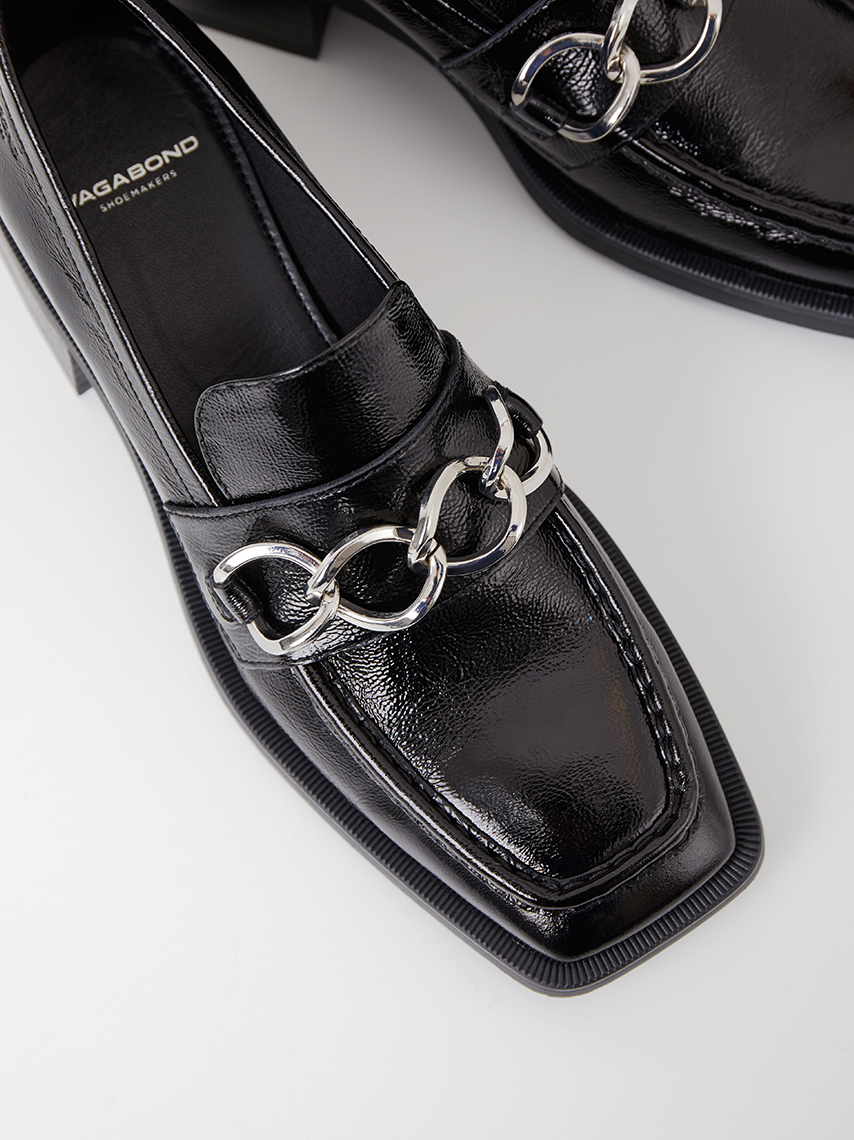 A detail view of Blanca loafers in polished black leather, with sharp heels and chain details in silver